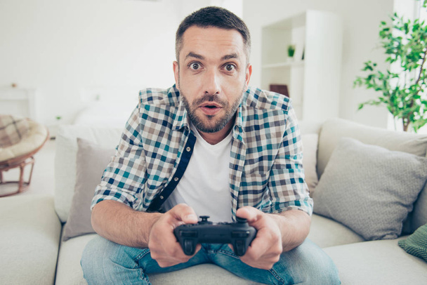 Close up photo of focused handsome mature guy hold hand have play station game pad dressed plaid denim jeans sit divan bearded amazed big apartment - Foto, imagen