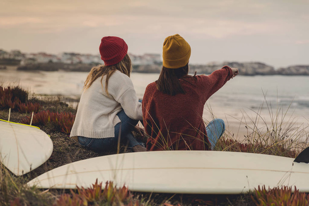 Two best friends sitting near the coastline with her surfboards while looking to the ocean - Foto, afbeelding