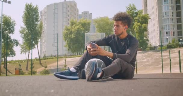 Retrato de primer plano del joven fuerte jugador de baloncesto afroamericano que escucha música en vibraciones en su teléfono sentado con una pelota al aire libre en la cancha
 - Metraje, vídeo