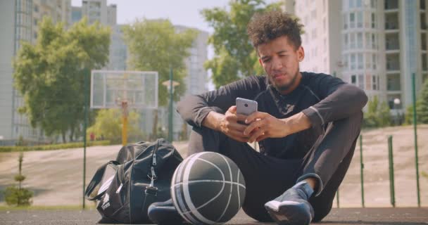 Closeup portrait of young strong african american male basketball player texting on the phone and looking at camera sitting with a ball outdoors on the court - Záběry, video