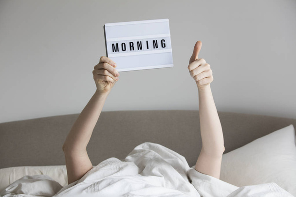 Femme au lit sous les draps tenant un panneau du matin
 - Photo, image