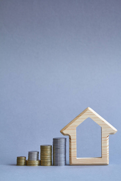 Dřevěná figurka v domě s několika sloupci mincí na šedém pozadí, koncept nákupu nebo pronájmu budovy, selektivní zaměření - Fotografie, Obrázek
