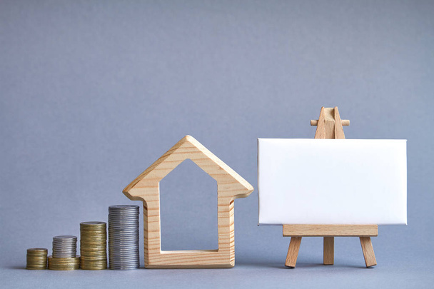 Wooden figure of house with several columns of coins nearby and white board on miniature easel on gray background, the concept of buying or renting a building, selective focus - Photo, Image