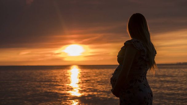 Jeune femme enceinte avec un gros ventre regarde le coucher de soleil majestueux sur la mer. En prévision d'un enfant
 - Photo, image