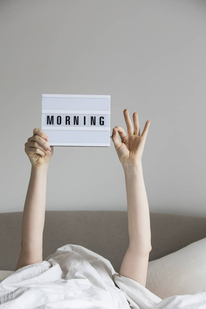 Female in bed under the sheets holding up a morning sign - Foto, imagen
