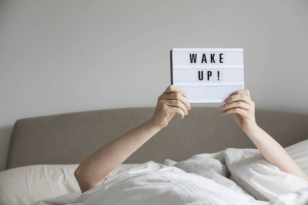 Female in bed under the sheets holding up a wake up sign - Foto, Imagem