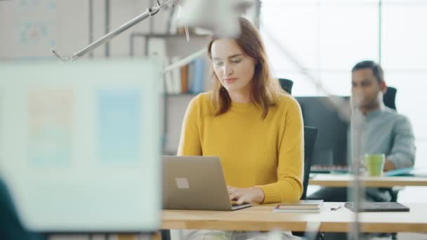 Beautiful and Creative Young Woman Sitting at Her Desk Using Laptop Computer. In the Background Bright Office where Diverse Team of Young Professionals Work - Footage, Video
