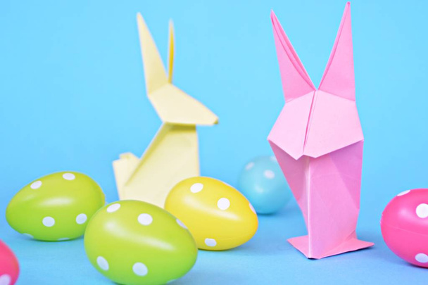 Origami bunnies folded from pastel paper stand on a blue base with dotted easter eggs on the outside - concept with origami for Easter with strong colors - Foto, afbeelding