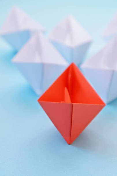 A red self-folded paper boat lies on a blue surface, followed by many white boats - concept symbolizing leadership - Photo, Image