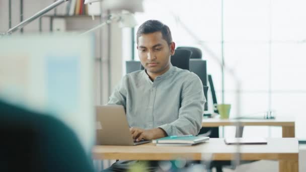 Smart and Handsome Indian Information Technology Specialist Sitting at His Desk works on a Laptop. In the Background Modern Office with Diverse Team of Young Professionals Working - Metraje, vídeo