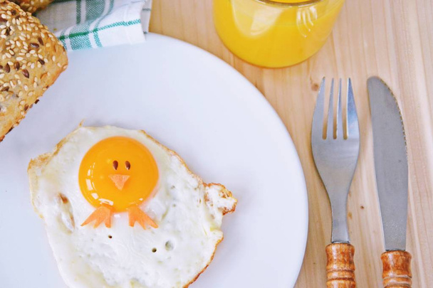 Een gebakken ei ligt op een bord, naast het zijn broodjes en bestek. De eierdooier van het gebakken ijs ziet eruit als een kuiken, met twee korrels van de ogen en stukjes wortel als mond en voeten - Foto, afbeelding