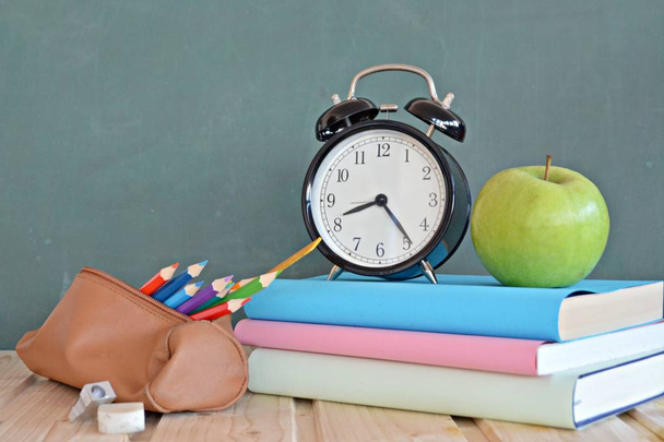 An alarm clock stands in front of a blackboard with colorful books and a green apple - concept with space for text or other elements on the subject of school and time management in elementary school - Photo, Image