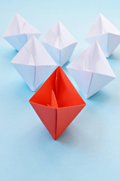 A red self-folded paper boat lies on a blue surface, followed by many white boats - concept symbolizing leadership - Zdjęcie, obraz