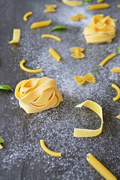 The word noodle was written in a flour-polluted area, along with various types of pasta such as penne and farfalle and basil leaves on a dark marble surface - Photo, Image