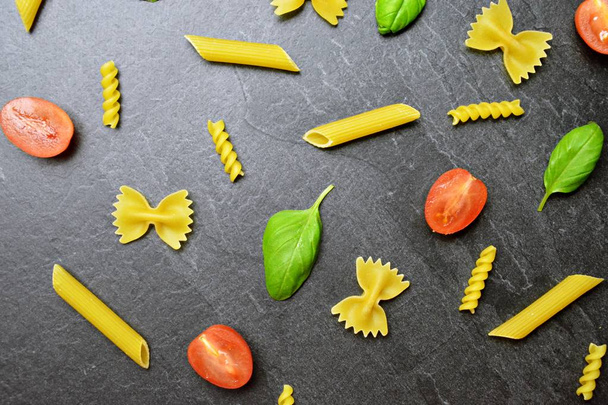 Various types of pasta such as rigatoni or farfalle lie on a dark marble surface along with half tomatoes and basil leaves - concept and background for fresh pasta  - Photo, Image
