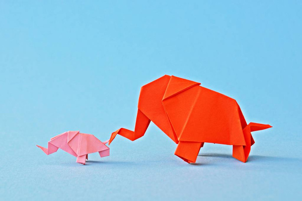 Mały i duży słoń origami złożony z czerwonego i różowego papieru stoją na monochromatycznej powierzchni przed odłamkiem lustro, które wygląda jak dwa słonie stoją przed otworem wody  - Zdjęcie, obraz