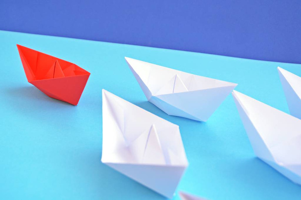 A red self-folded paper boat lies on a blue surface, followed by many white boats - concept symbolizing leadership - 写真・画像