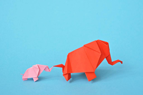 Dos elefantes plegados en papel en soporte rosa y rojo sobre un fondo azul - concepto con un pequeño elefante bebé y un elefante madre plegado de acuerdo con la técnica de origami
 - Foto, Imagen