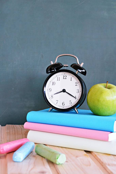 目覚まし時計は、カラフルな本と緑のリンゴと黒板の前に立っている - 小学校の学校や時間管理の主題にテキストや他の要素のためのスペースを持つコンセプト - 写真・画像