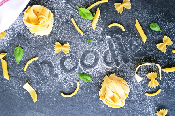 The word noodle was written in a flour-polluted area, along with various types of pasta such as penne and farfalle and basil leaves on a dark marble surface - Photo, Image