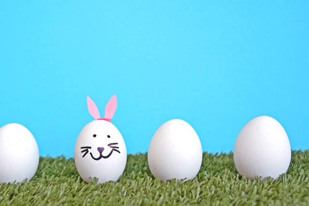 яйца, раскрашенные лицом зайца и приклеенные к кроличьим ушам, лежат на травяном поле перед деревянным фоном с местом для текста или других элементов
 - Фото, изображение