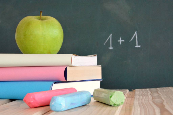 Three books with different colored covers lie on a wooden surface in front of a blackboard with an apple - concept for education and school with place for text or other elements - Photo, Image