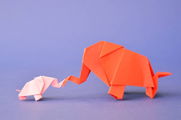 Mały i duży słoń origami złożony z czerwonego i różowego papieru stoją na monochromatycznej powierzchni przed odłamkiem lustro, które wygląda jak dwa słonie stoją przed otworem wody  - Zdjęcie, obraz