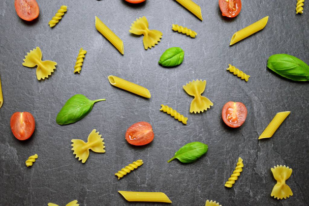 Various types of pasta such as rigatoni or farfalle lie on a dark marble surface along with half tomatoes and basil leaves - concept and background for fresh pasta  - Photo, Image