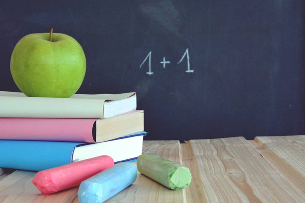 Τρία βιβλία με διαφορετικά χρωματιστά καλύμματα βρίσκονται σε μια ξύλινη επιφάνεια μπροστά από έναν πίνακα με ένα μήλο-concept για την εκπαίδευση και το σχολείο με τόπο για κείμενο ή άλλα στοιχεία - Φωτογραφία, εικόνα