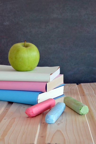 drei Bücher mit unterschiedlich farbigen Einbänden liegen auf einer Holzfläche vor einer Tafel mit einem Apfel - Konzept für Bildung und Schule mit Platz für Text oder andere Elemente - Foto, Bild
