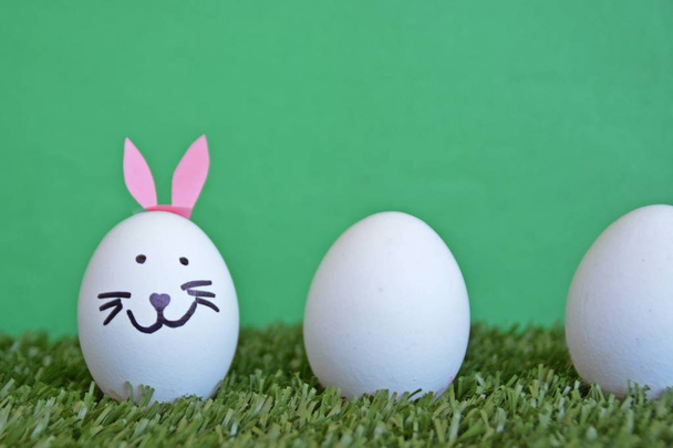 eggs painted with the face of a hare and glued to rabbit ears lie on a grass field in front of a wooden background with space for text or other elements - Photo, Image