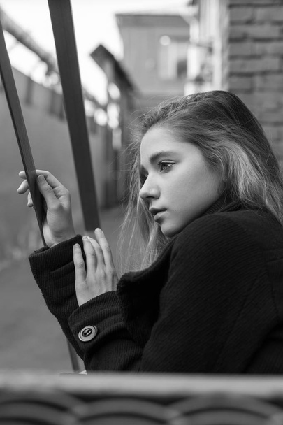 Λυπημένη νεαρή κοπέλα με μαύρο παλτό κάθεται στις σκάλες με vintage αστικό υπόβαθρο. Έννοια της μοναξιάς. - Φωτογραφία, εικόνα