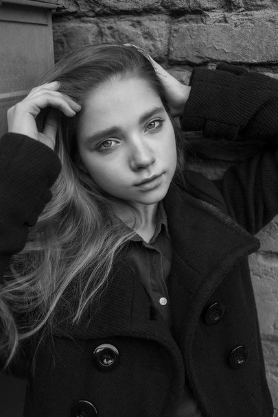 Siyah ceketli, uzun saçlı üzgün şehvetli kız duvarın yanında oturuyor. Melankoli kavramı. Siyah ve beyaz resim - Fotoğraf, Görsel