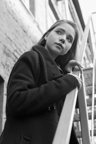 Young girl in black coat standing on stairs with brick wall on background. Concept of loneliness. Black and white image - Fotografie, Obrázek