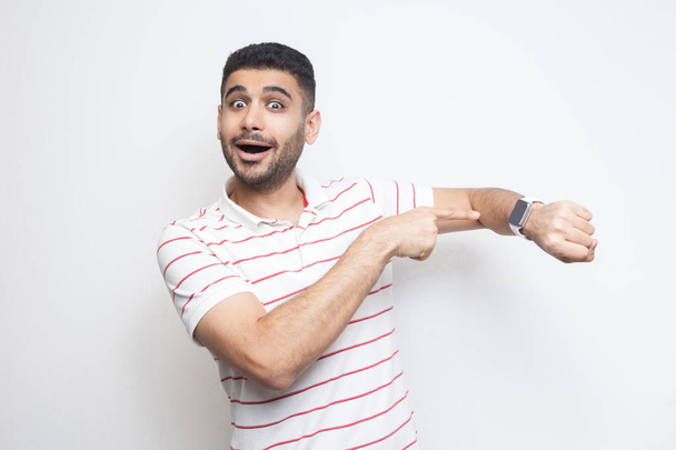  amazed funny bearded young man in striped t-shirt pointing at smart watch and looking at camera with surprised face isolated on white background - Photo, Image