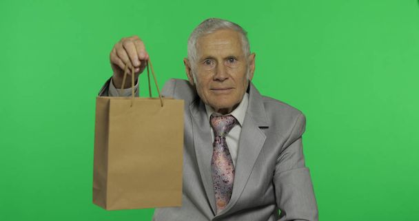 Γέρος με τσάντα για ψώνια. Κοιτάζει μέσα στην κάμερα και τεντώνει το δώρο - Φωτογραφία, εικόνα