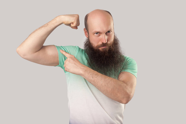 serious middle aged bald man with long beard in light green t-shirt pointing his bicep and looking at camera with haughty face on grey background. - Foto, Bild