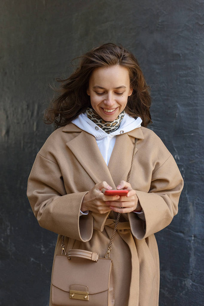 Pouliční portrét mladé usměvavé ženy, oblečené v béžové kabátě pomocí inteligentního telefonu venku. Moderní internetová komunikace - Fotografie, Obrázek