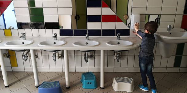 Pequeño chico caucásico se lava las manos en el baño de niños en el jardín de infantes, de pie con la espalda a la cámara
 - Foto, Imagen