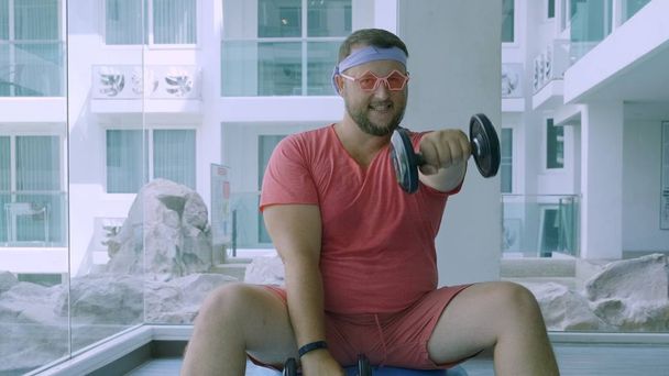 Παιχνιδιάρικο λίπος άνθρωπος σε ένα ροζ T-shirt και ροζ γυαλιά ασχολούνται με το γυμναστήριο με βαράλια και μια μπάλα Fit στο γυμναστήριο - Φωτογραφία, εικόνα