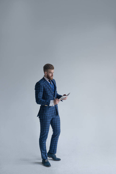 Volledige lengte shot van jonge bebaarde stijlvolle zakenman leider binnenshuis met driedelige pak met horloge houden krant en kijken weg op grijs - Foto, afbeelding