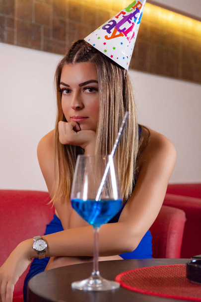 Młoda śliczna dziewczyna z czapką urodzinowa na głowie siedzi w restauracji i picia Błękitnej Laguny koktajl i niewyraźne tło. Zbliżenie, selektywne skupienie - Zdjęcie, obraz