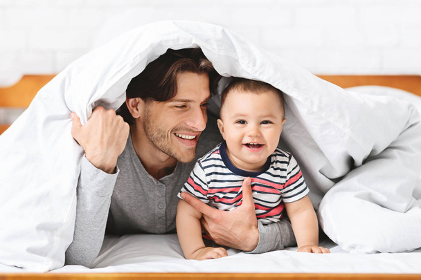 Juguetón padre escondido bajo manta con bebé hijo
 - Foto, imagen