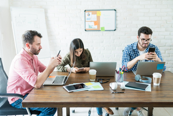Empresários sentados em torno da mesa de conferências enquanto socializam em telefones celulares no escritório
 - Foto, Imagem