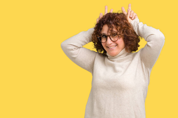 Mooie midden ager senior vrouw die coltrui trui en bril dragen over geïsoleerde achtergrond die zich voordeed grappig en gek met vingers op hoofd als bunny oren, lacht vrolijk - Foto, afbeelding