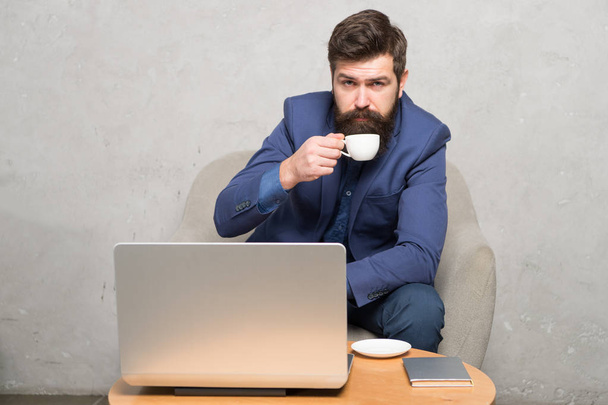 In mobile style. Bearded man enjoying his hot drink and mobile internet. Businessman with mobile device drinking tea or coffee at workplace. Hipster in formalwear working at laptop in mobile office - 写真・画像