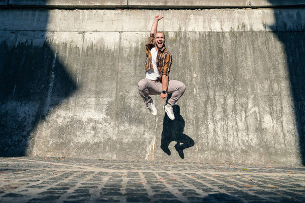 Rock Your Life. Jonge goede uitziende vette bebaarde man springt buiten tegen grijze moderne loft muur met zijn hand omhoog. gelukkige gezichtsuitdrukking. dragen van geel overhemd en jeans. - Foto, afbeelding