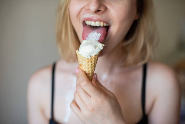 Sexy blonde licks a waffle cone. Ice cream is melting, erotically draining through the body of an attractive woman. Beautiful girl in black underwear smeared with ice cream. Close-up - Zdjęcie, obraz