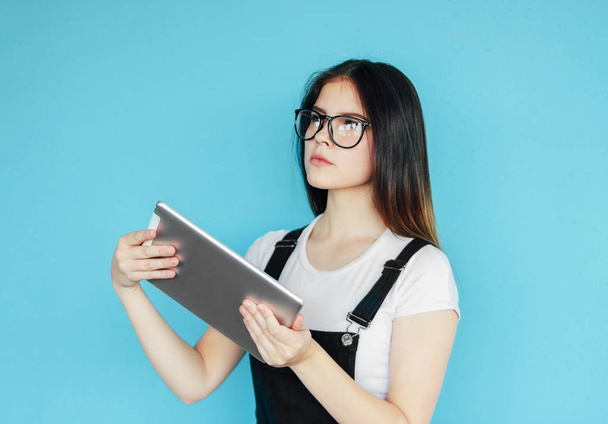 Серйозна красива брюнетка дівчина-підліток в окулярах і біла футболка тримає планшет в руках, ізольована на синьому фоні
 - Фото, зображення