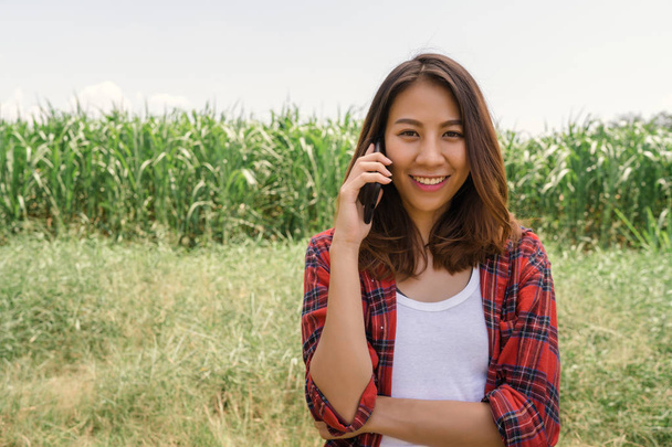 Alegre asiática agricultora y empresaria posando en la cosecha de maíz y sonriendo usando el teléfono móvil o la aplicación para teléfonos inteligentes, en cámara, agricultura y concepto de cultivo
. - Foto, imagen